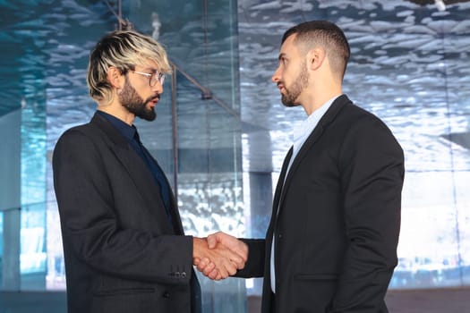 Two professional businessmen in suits shaking hands inside a modern glass office. Business deal, agreement, or partnership in a corporate indoor setting with contemporary design.