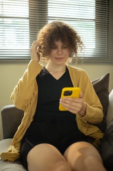 Relaxed woman sitting on sofa with headphones and using smartphone.