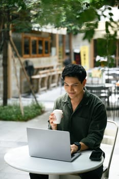 Adult man enjoying coffee while working remotely on his laptop at a modern outdoor cafe.