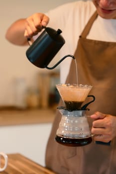 Young man in apron brewing coffee using the pour-over method in warm kitchen.