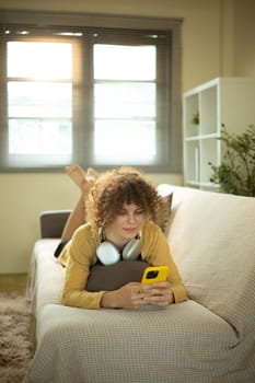 Happy woman with curly hair using mobile phone while lying comfortably on the couch.