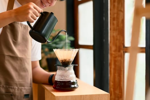 Young man in apron brewing coffee using the pour-over method in warm kitchen.