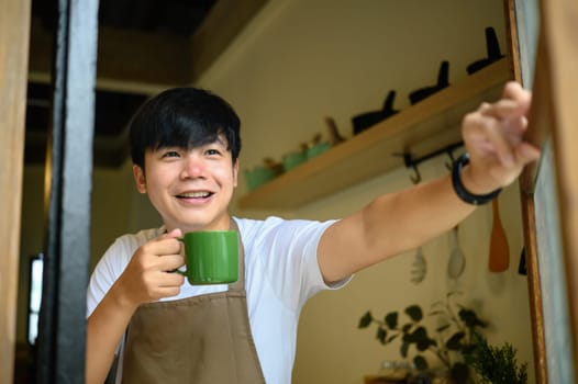 Young man holding a coffee cup smiling warmly as he opens a window in a cozy kitchen.