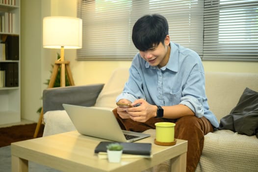 Smiling man using smartphone with laptop on coffee table in living room.
