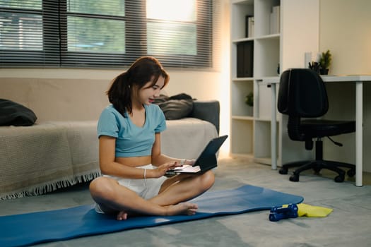 Asian woman in sportswear checking her fitness routine online sitting on mat in living room.