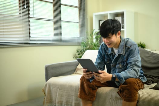 Asian male in denim jacket using digital tablet on couch in a bright living room.