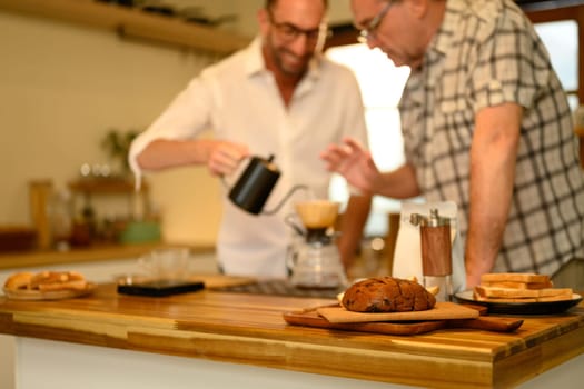 Sliced loaf of bread on a wooden countertop with father and son preparing coffee in background.