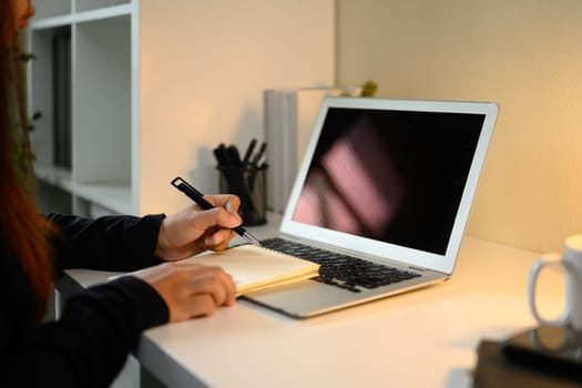 Close up of woman writing in notebook next to blank laptop screen on modern workspace.