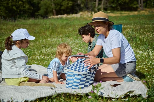 A mother and her children engage in a delightful summer picnic on a flower-filled meadow in the mountains. The family shares quality time outdoors under bright sunshine, embracing nature.