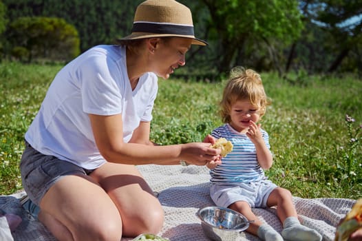 A woman and her young child share a joyful picnic scene outdoors. The warm and sunny day enhances the serene mountain backdrop, emphasizing family bonding and love for nature.