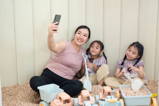 A joyful mother takes a selfie with her two daughters while playing with toys in a cozy playroom, highlighting family connection and fun.