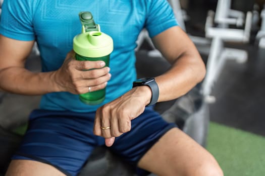 A fit man holding a green water bottle while checking his smartwatch during a workout session at the gym. Focus on fitness and hydration.