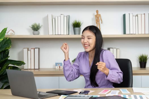 A joyful graphic designer celebrates a successful project at her workspace, surrounded by design materials and a laptop, showcasing enthusiasm and creativity.