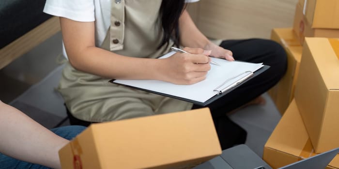 A woman writes notes while her partner organizes packages in their home office, showcasing their teamwork in running an online business.