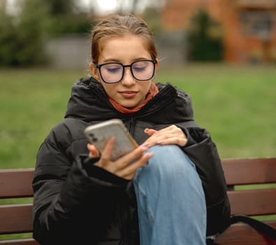 In Spring, A Girl In Glasses Sits On A Bench With A Smartphone