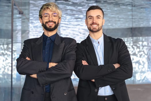 Two confident male professionals in suits standing with crossed arms, smiling at the camera inside a modern office. Corporate success, teamwork, and leadership in business environment.