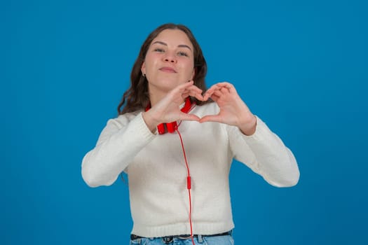 Happy young woman in white sweater and red headphones smiling while making a heart shape with her hands against a blue background, representing kindness, connection, love, and positive emotions.