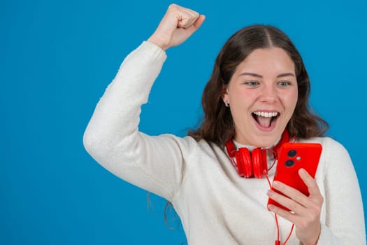 Happy young woman wearing red headphones around her neck, clenching her fist in excitement while looking at her smartphone. Her joyful reaction stands out against the blue background.Copy space