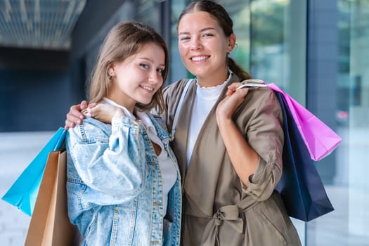 Two happy caucasian women holding colorful shopping bags and smiling after a successful day at the mall. They stand outside a modern building, enjoying their time together with style and joy.
