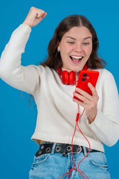 Happy young woman wearing red headphones around her neck, clenching her fist in excitement while looking at her smartphone. Her joyful reaction stands out against the blue background.Vertical