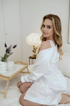 Young blonde in a white robe smiling and having breakfast in a white bedroom on the bed. table in the bed. concept of the morning of the bride, breakfast in bed, morning coffee.