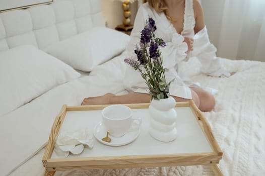 Cropped Young girl in white silk robe having breakfast in white bedroom on bed. table in bed. concept of bride morning, breakfast in bed, morning coffee. White dishes on table and lavender in vase