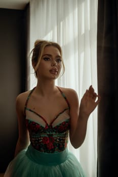 young slender blonde looks at herself in the mirror room. Turquoise dress with a corset, with red embroidered flowers, open shoulders on straps against the background of the window