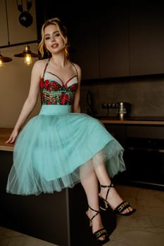 Smiling young blonde in a turquoise dress poses in the kitchen. SheFluffy skirt and corset sits on the table and smiles playfully. Looking at the camera. Minimalistic interior in modern style.