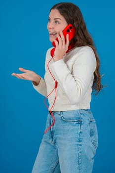 Vertical image of a cheerful young woman talking on a red smartphone with matching headphones. She laughs with hand on chest, standing against a bright blue background in a casual moment.