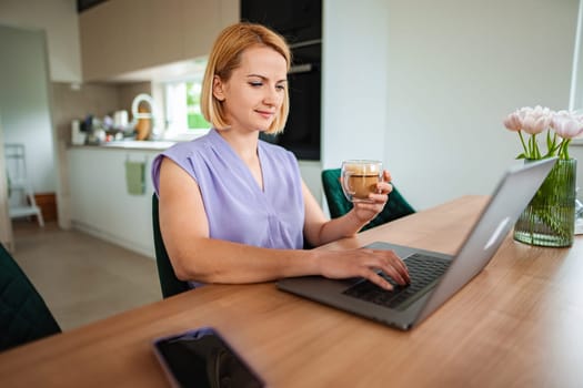 Woman enjoys coffee and works on laptop in cozy kitchen.
