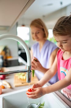 Young child washes vegetables while adult prepares food.