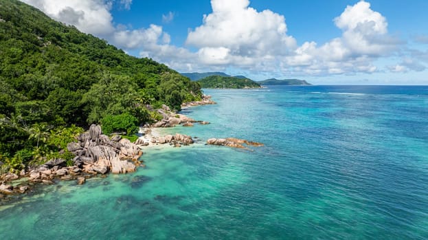 A scenic tropical coastline featuring large rocks, lush green vegetation, and beach. Praslin, Seychelles.