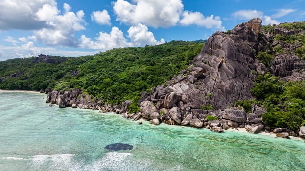 Steep cliffside covered with lush green foliage, overlooking turquoise waters with gentle waves under a clear sky. Seychelles, La Digue.