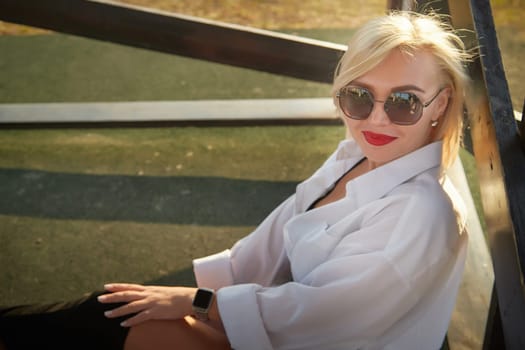 A woman poses comfortably outdoors wearing a stylish outfit and sunglasses, enjoying the warm sunlight in the late afternoon.