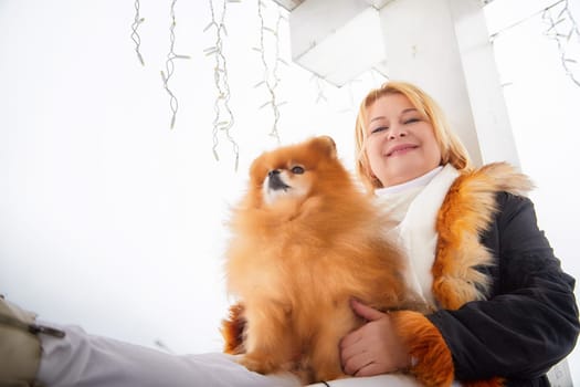 A middle-aged blonde woman happily pets her fluffy red Pomeranian dog while enjoying the snowy winter scenery.