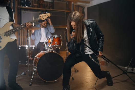 Expressive rocker singing song in microphone while his band playing heavy metal on drum and electric bass guitar. Young guys at rehearsal repetition in home studio