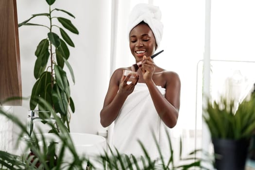 A woman with a towel on her head smiles while applying cream from a jar in a sunlit, stylish bathroom filled with plants. The atmosphere is calm and refreshing.