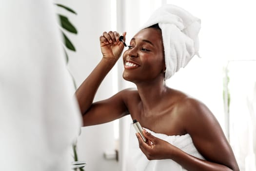 A woman with a towel wrapped around her hair and body smiles while applying mascara in a well-lit bathroom. She enjoys her beauty routine, showcasing self-care and confidence.