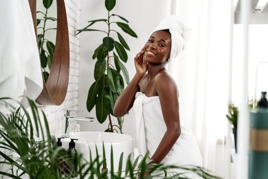A woman with a towel wrapped around her body and a towel on her head smiles while applying skincare in a bright, stylish bathroom filled with plants and natural decor.