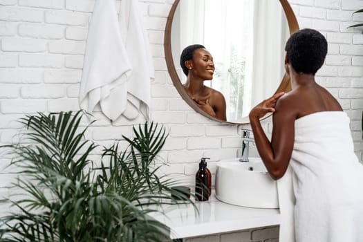 In a bright and modern bathroom, a woman wrapped in a towel smiles at her reflection while applying skincare. Lush plants and a round mirror enhance the calming atmosphere.