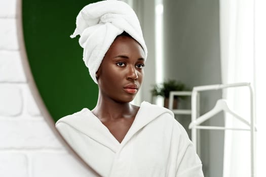 A woman with a towel wrapped around her hair looks thoughtfully into a mirror. She wears a plush white robe, and the bathroom features soft lighting and minimal decor.