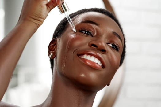 In a well-lit bathroom, a woman with short hair applies a serum to her face using a dropper, displaying joy and confidence during her skincare routine.