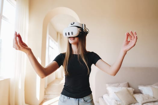 A young woman wears a virtual reality headset and moves her arms in an immersive experience. The bright room features soft furnishings and large windows, creating an inviting atmosphere.