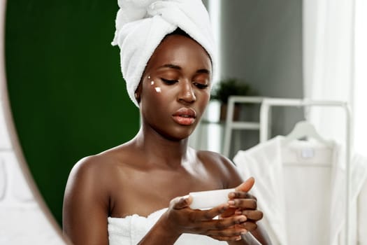 A woman with natural skin tones and a towel wrapped around her head is applying a skincare product while looking at her reflection in the mirror. The bright bathroom setting enhances her routine.