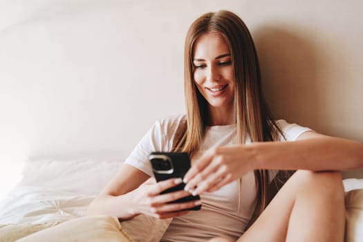 A young woman sits comfortably on her bed in a bright bedroom, smiling at her smartphone. She wears a casual white t-shirt and looks relaxed, enjoying leisure time in the morning.