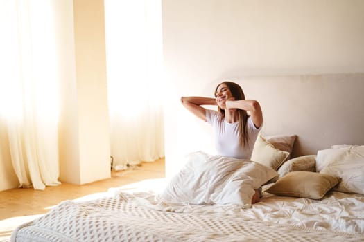 A woman wakes up in a cozy bedroom, stretching with joy as the morning sunlight fills the space, surrounded by fluffy pillows and warm blankets.