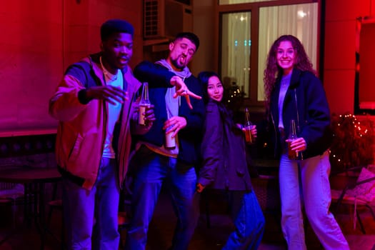 Four friends enjoy a fun evening together in a cozy indoor space, illuminated by vibrant colorful lights while holding drinks and posing playfully.
