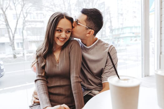 A young couple relaxes at a cafe, exchanging affectionate gestures while seated at a table. They share laughter and warmth amidst a bustling urban backdrop on a sunny day.