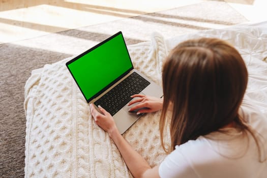 A young individual is focused on a laptop with a green screen while sitting on a comfortable bed covered with a knitted blanket. Sunlight fills the room.