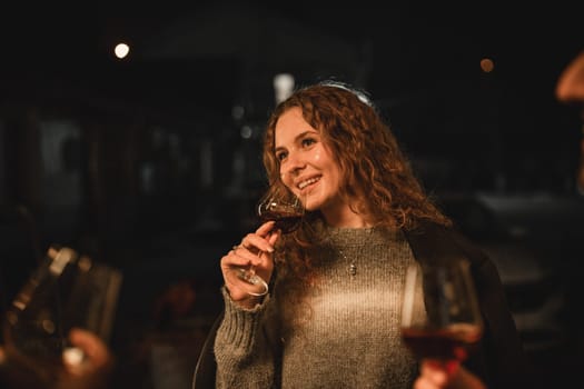 A woman with curly hair enjoys a glass of wine during an evening gathering. Friends surround her in a warm and lively atmosphere under the stars, sharing laughter and conversation.